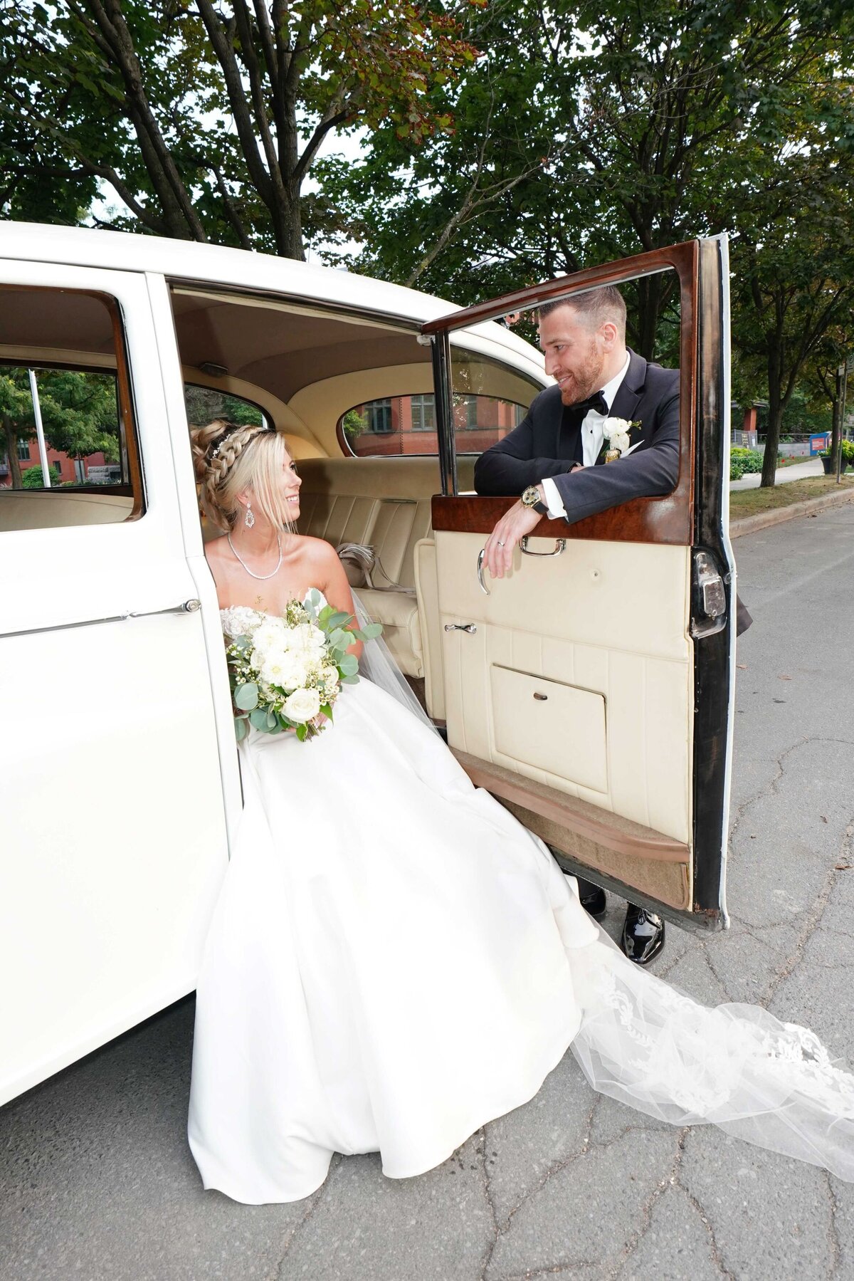 The bride sits inside a car, while the groom leans on the car door, gazing at her with affection. The image captures a tender, candid moment between the couple, set against the backdrop of their vehicle.