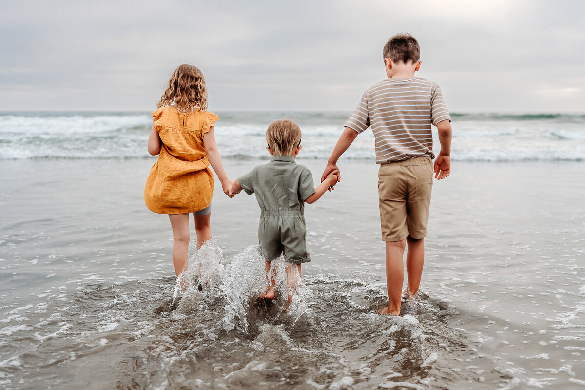 Three siblings holding hands and walking into the ocean waves at Del Mar beach during a San Diego family photography session.
