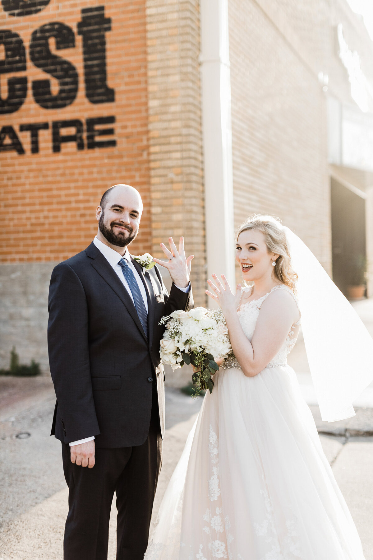 A portrait of a bride and groom playfully showing off their wedding rings after their ceremony in Fort Worth, Texas. The bride is on the right and is wearing a long, flowing white dress and a white veil while holding a bouquet of white flowers. The groom is on the left and is wearing a black suit with a blue tie and boutonniere.