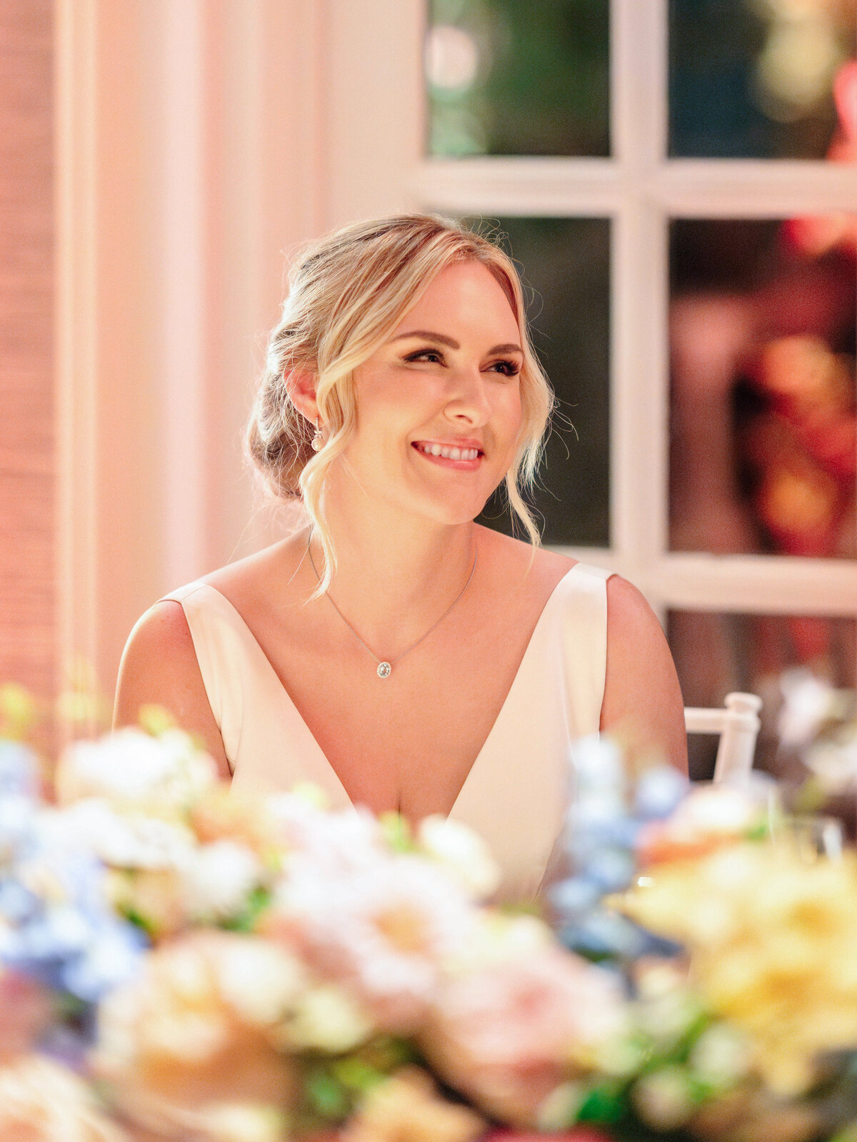 A woman with blonde hair tied up is smiling while sitting at a table. She is wearing a white dress and a necklace. The background shows a window and blurred greenery. There are colorful flowers in the foreground.