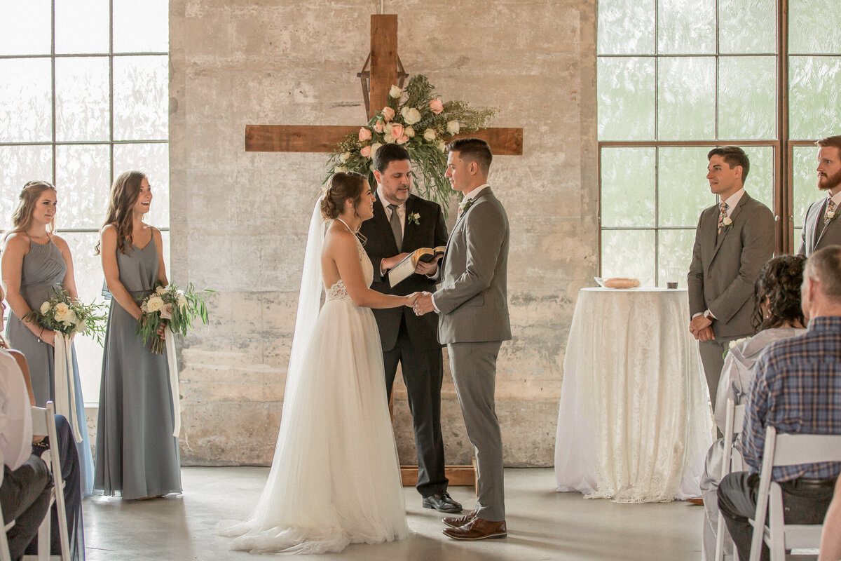 the bride and groom face each other in front of their officiant and a cross decorated with pink and white florals.  Their wedding party looks on, smiling.