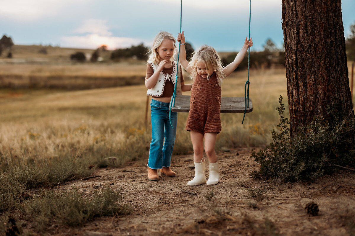 two young sisters playing on an outdoor wooden swing attached to a tree