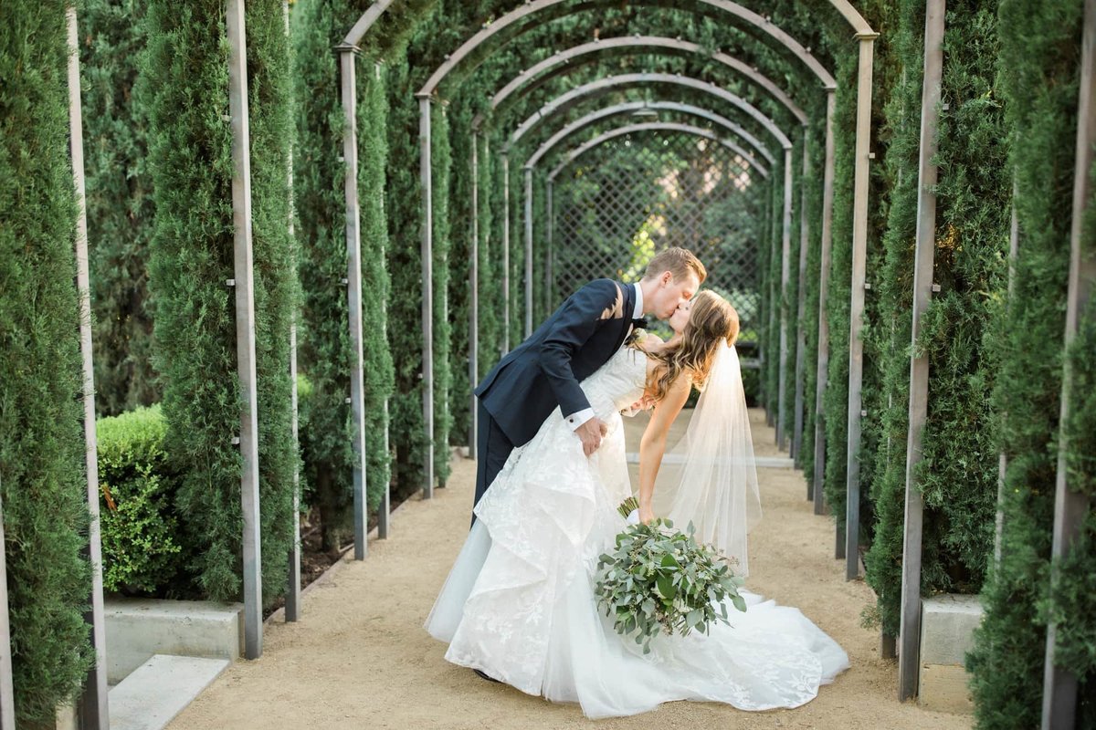 Groom dips and kisses his Bride as they walk under a tunnel covered by foliage