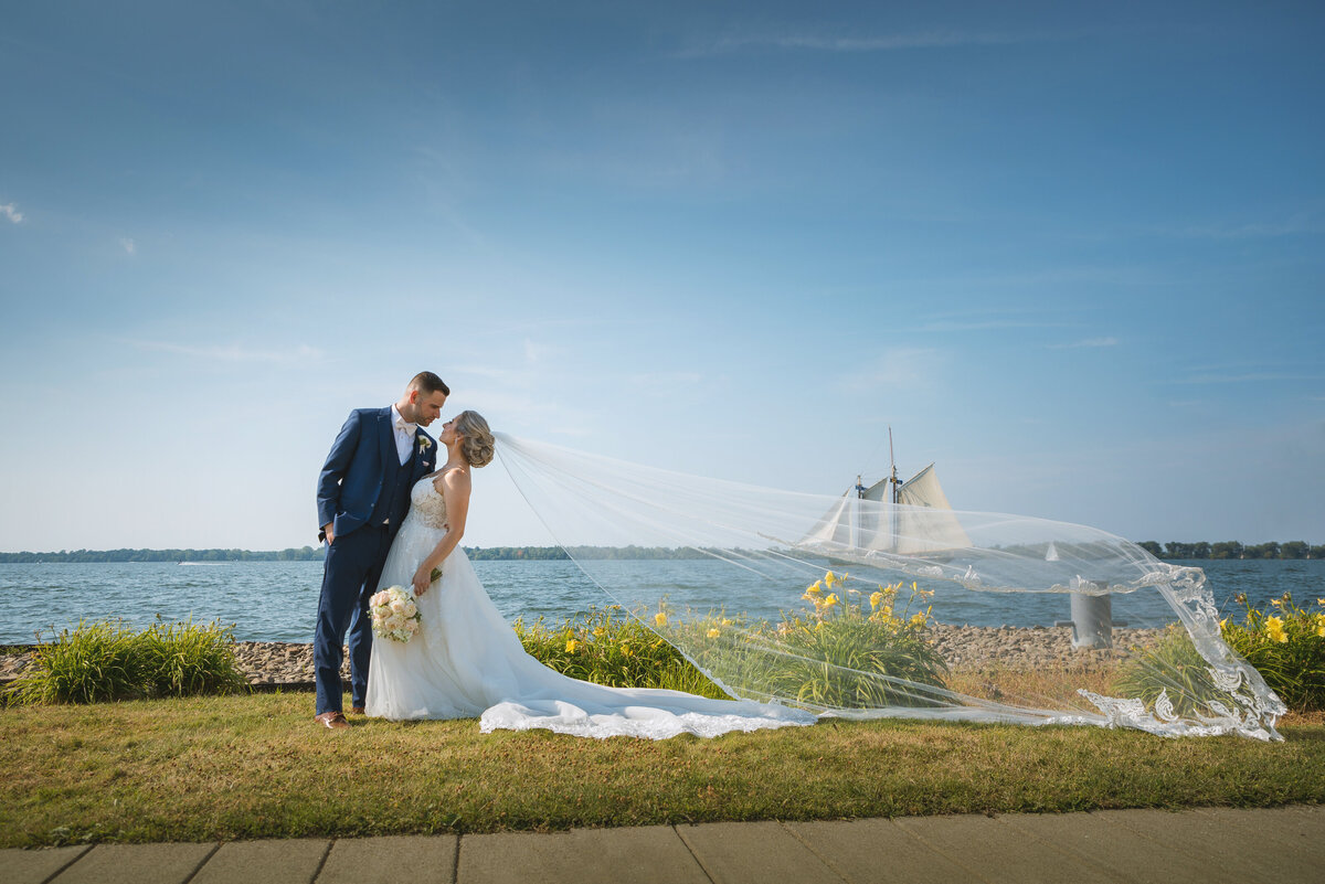 Bride and groom kissing with veil blowing and sailboat in background.