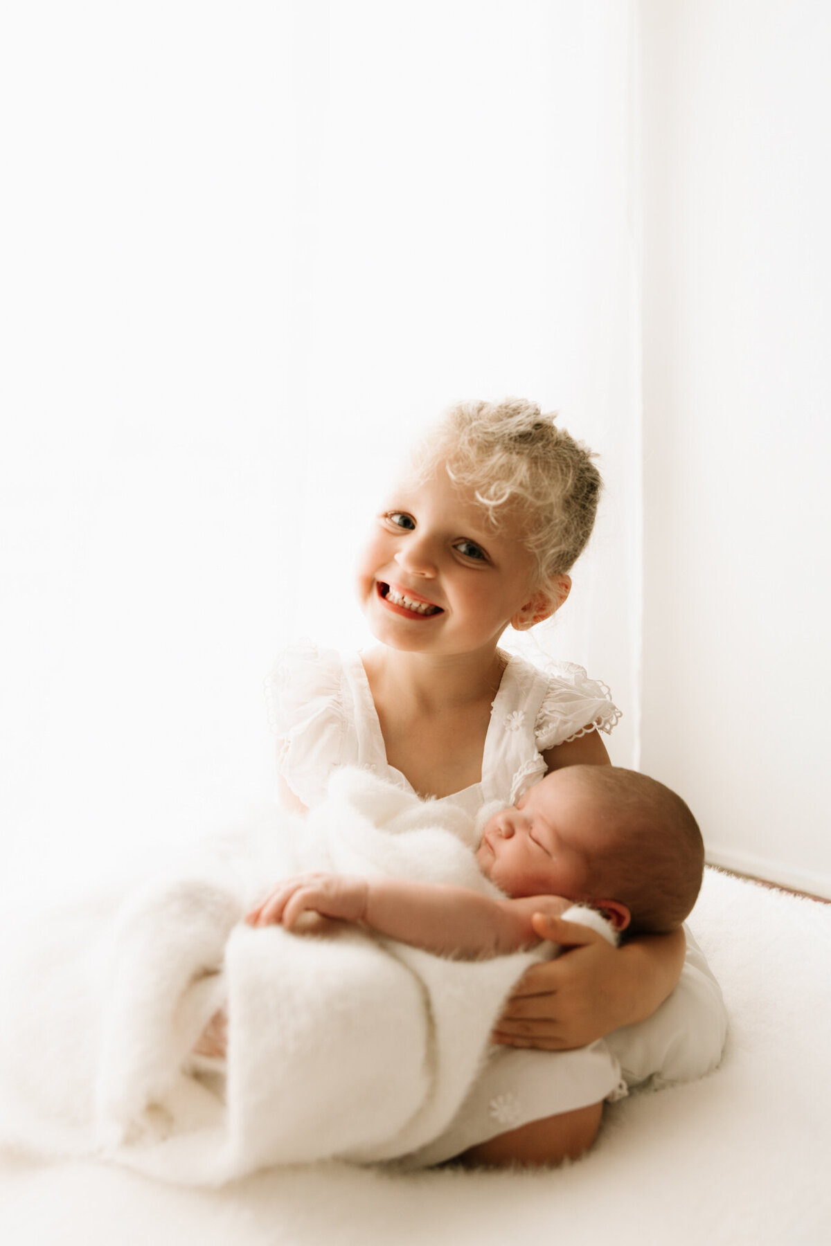 A joyful young girl with curly blonde hair and a bright smile lovingly holds her newborn sibling, who is swaddled in a soft white blanket. The siblings are seated on a plush white surface, bathed in soft, diffused light from sheer curtains behind them. The older sister wears a delicate white dress, adding to the timeless and heartwarming feel of this sibling portrait captured in a Sydney newborn photography studio.