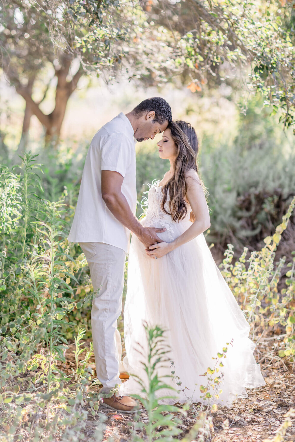 Husband and wife dressed in white putting their heads together and closing their eyes while cradling wife's baby bump in a field of green grass.