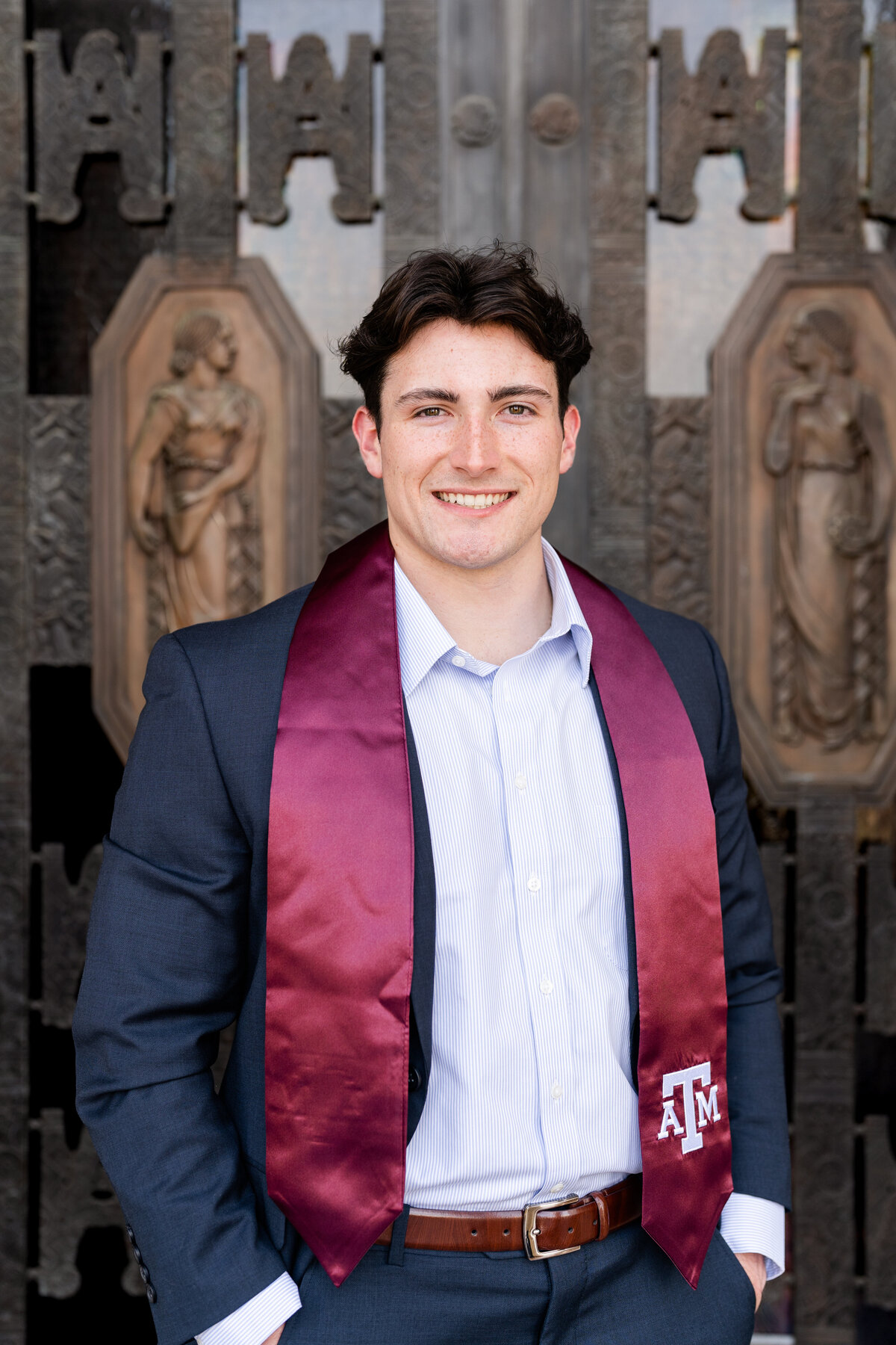 Texas A&M senior guy smiling with hands in pocket and wearing suit while standing in front of Administration Building doors