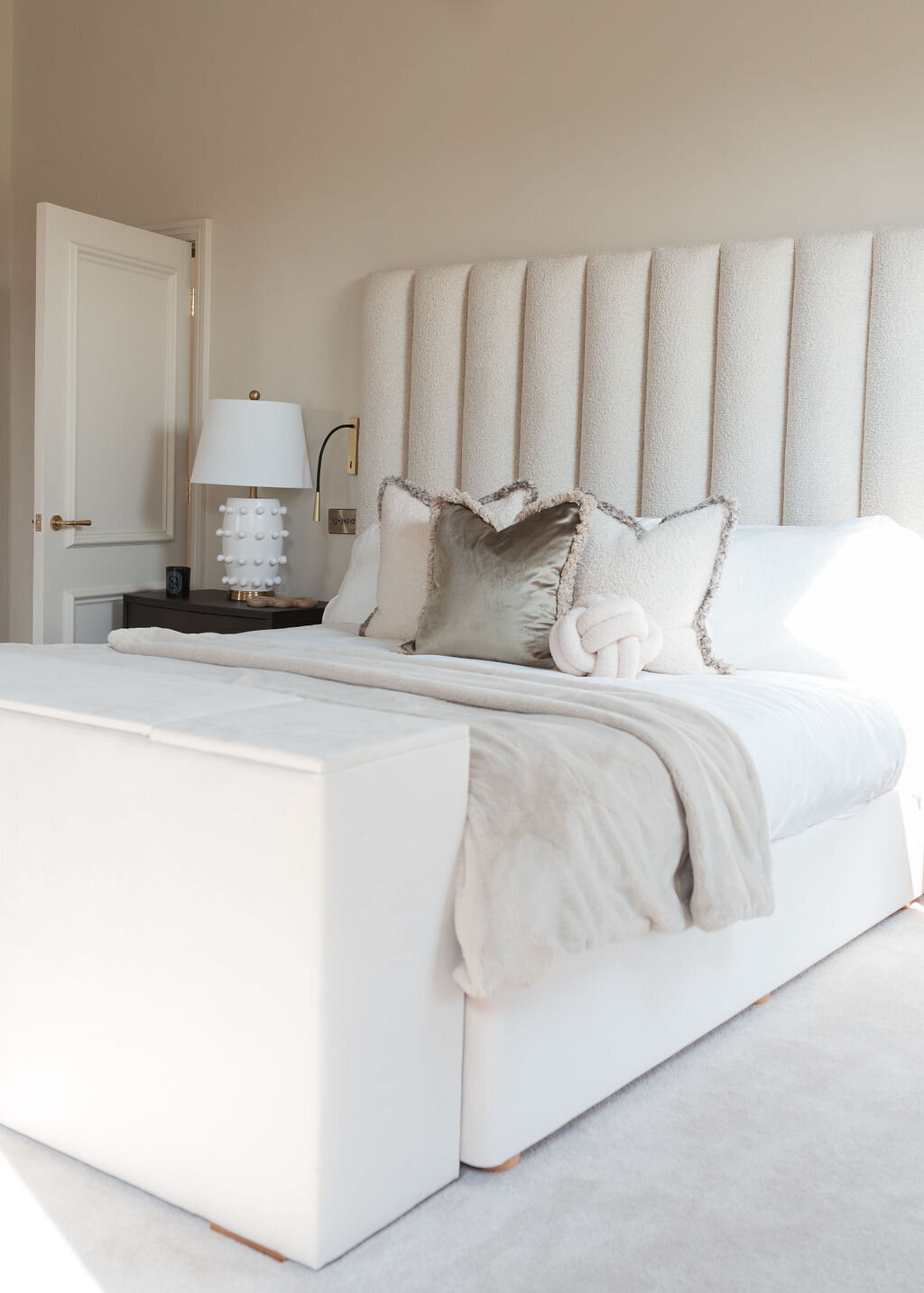 A well-lit bedroom featuring a large plush bed with a tall, tufted headboard. The room has a neutral color palette with white and beige bedding and some accent pillows.