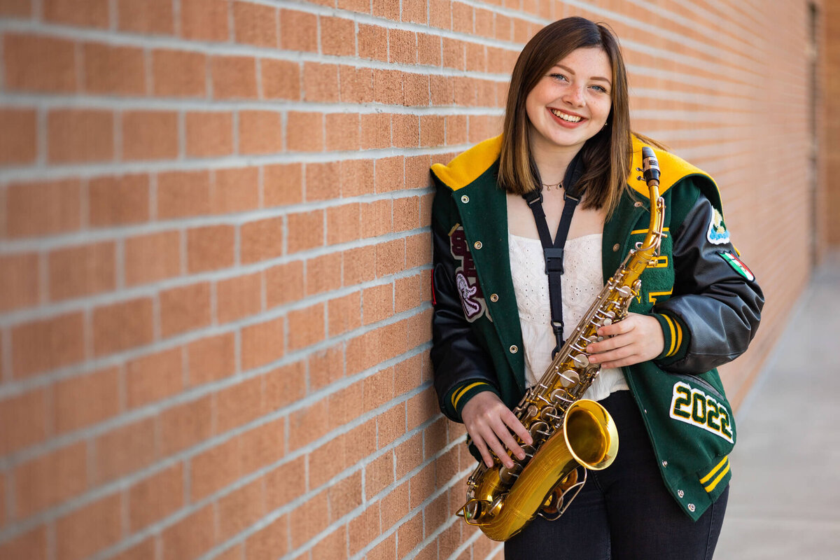 high school senior girl  wearing her letterman jacket  holding her saxophone leaning agains a brick wall