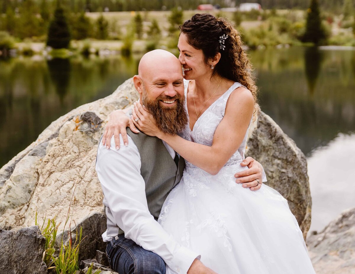 Bride embraces groom and laughs while sitting together on rocks beside a calm alpine lake in Colorado.