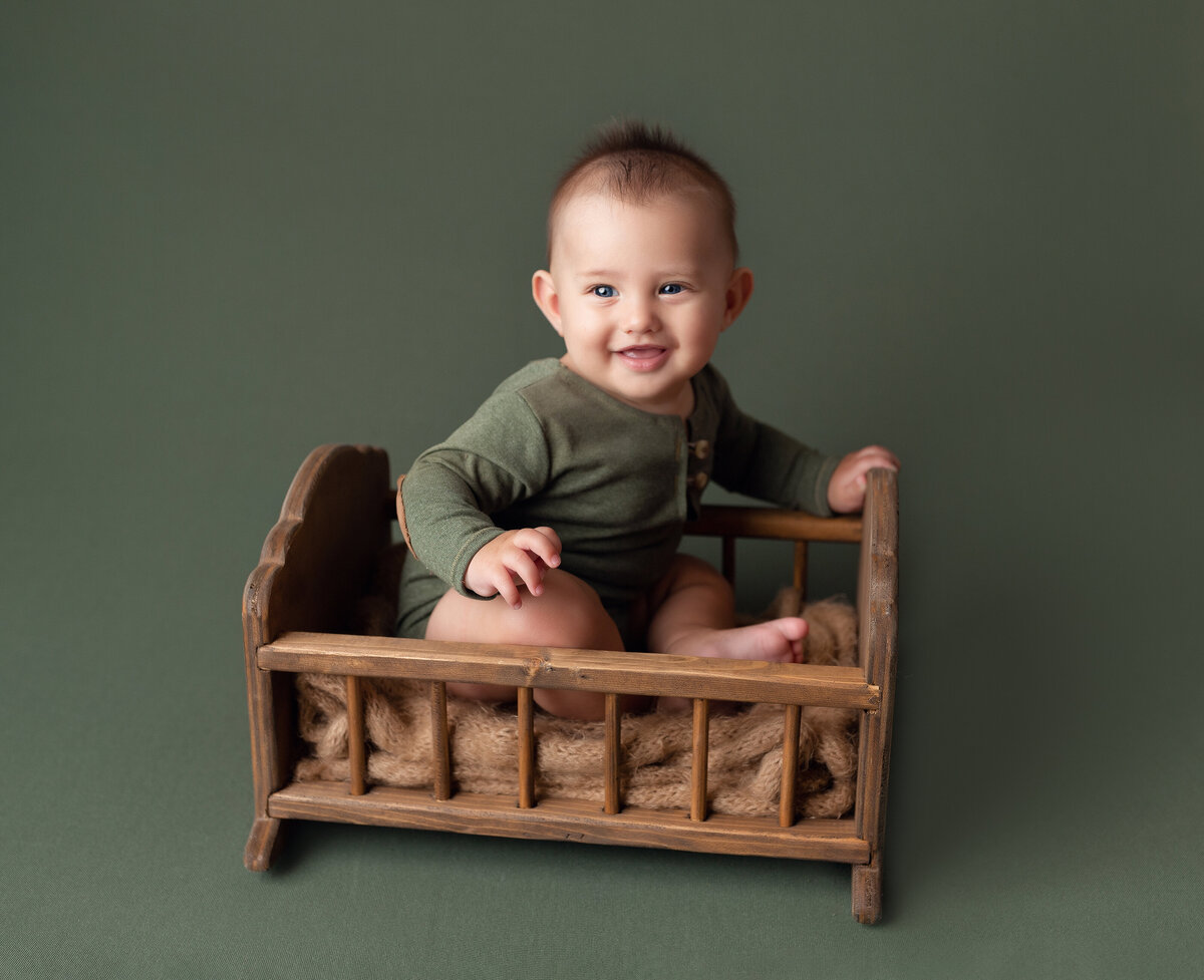 Baby boy 6-month milestone session at West Palm Beach and Boynton Beach photography studio. Baby is sitting in an antique miniature crib  on a green infinity backdrop. Baby is wearing a white onesie smiling up at the camera.