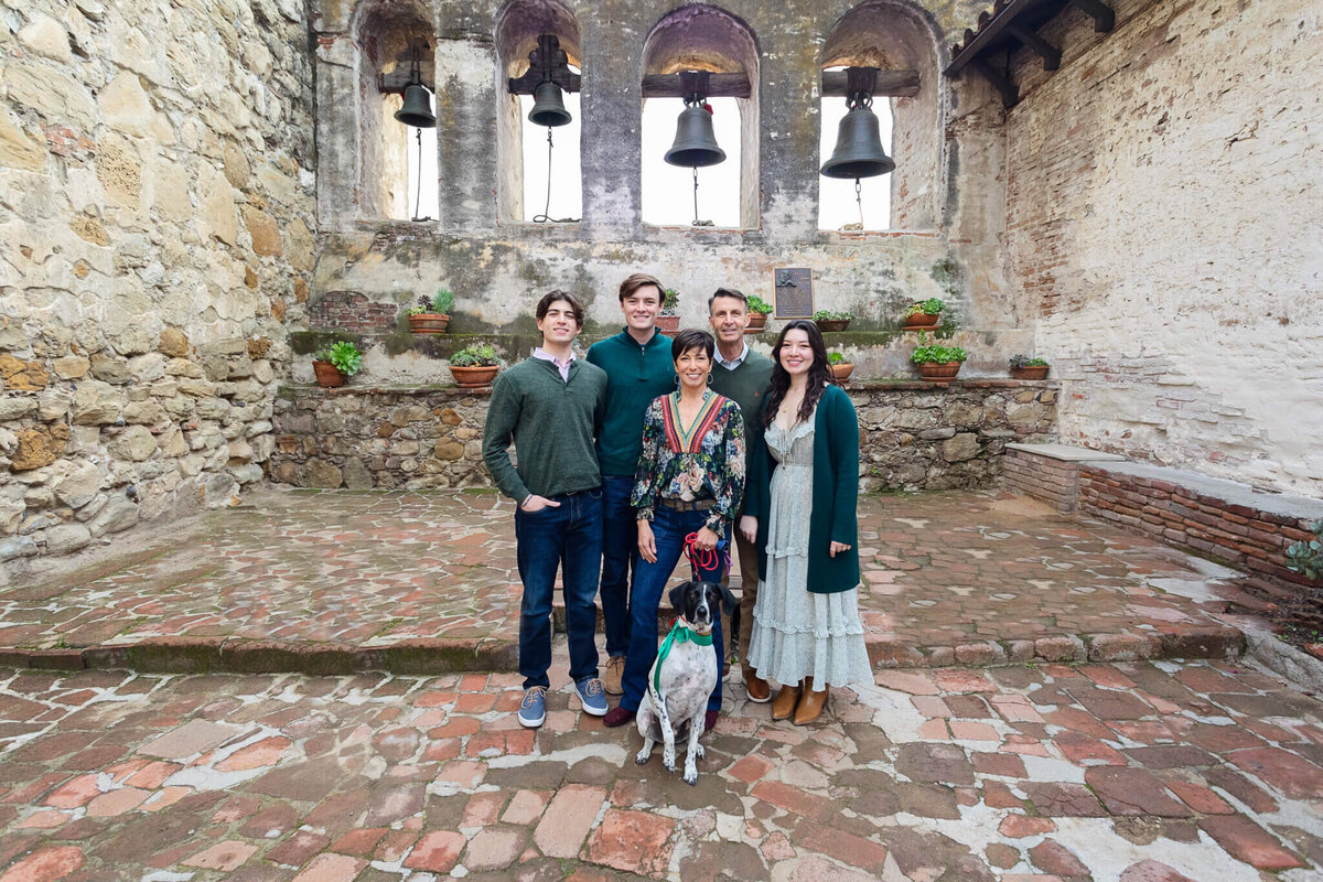 A family of five and their dog standing in a brick courtyard in front of archways with bells hanging in them.