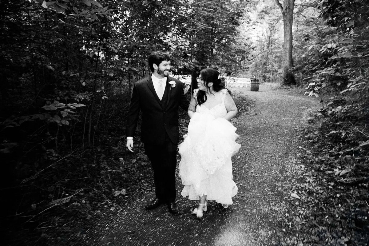 A bride and groom walk arm-in-arm down a forest path, smiling at each other. Captured by an NJ wedding photographer, the bride holds up her dress as they stroll, surrounded by dense trees and foliage.
