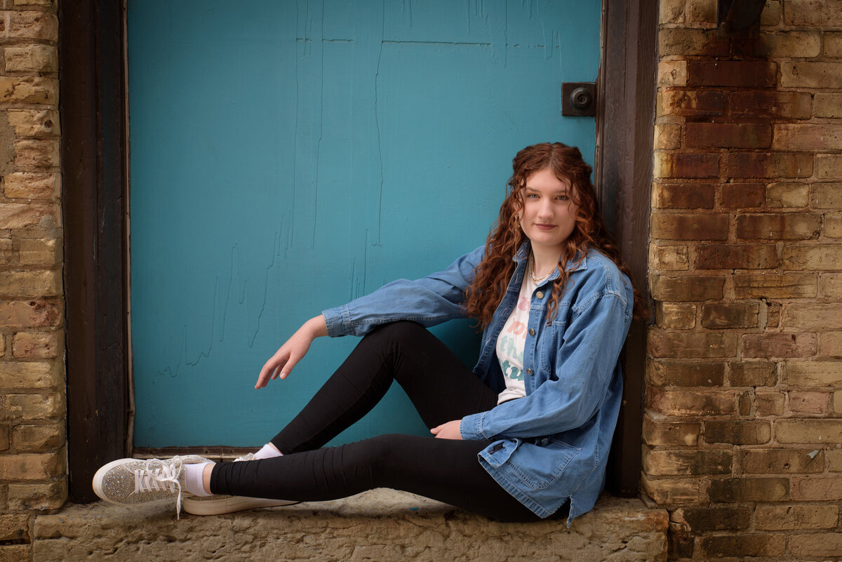 Green Bay East High School senior girl wearing a graphic t-shirt and leggings sitting in a doorway with a teal door and brick wall in an urban setting in downtown Green Bay, Wisconsin.