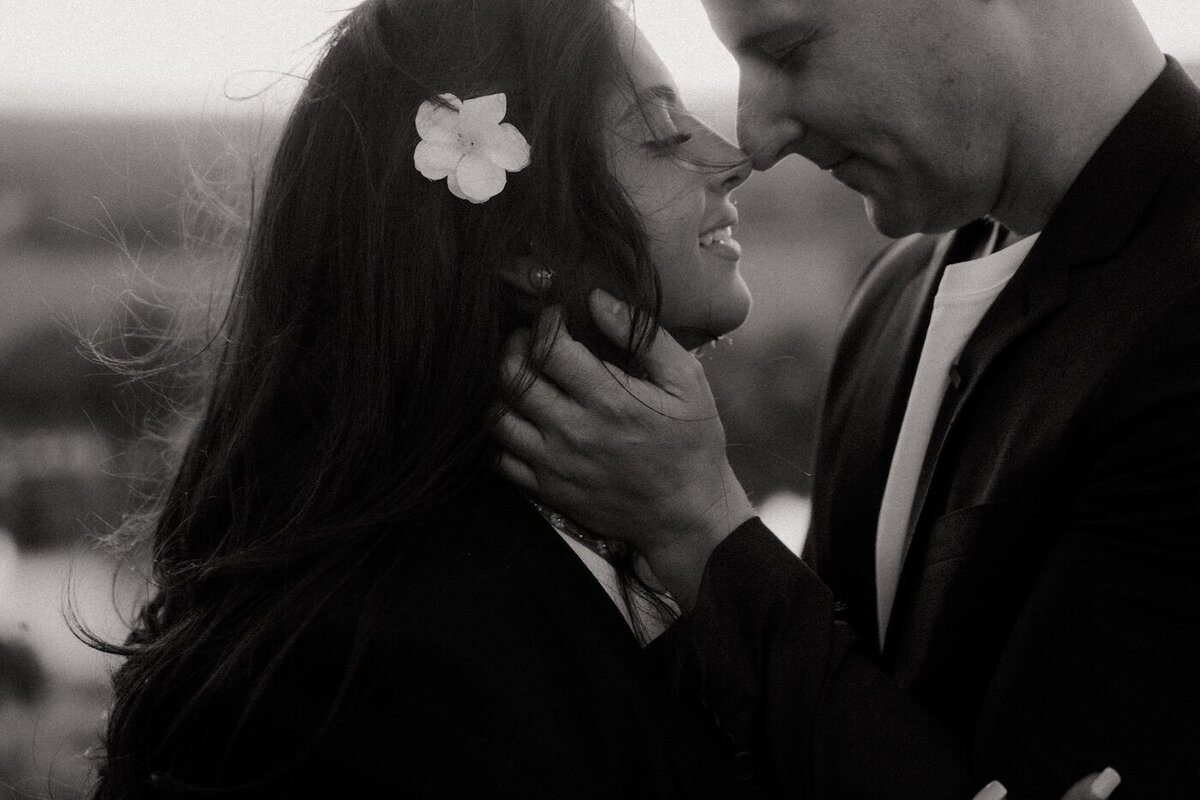 Couple intime, sourire complice, fleur blanche dans les cheveux de la femme. Portrait romantique en noir et blanc par Kamescop, séance engagement en extérieur
