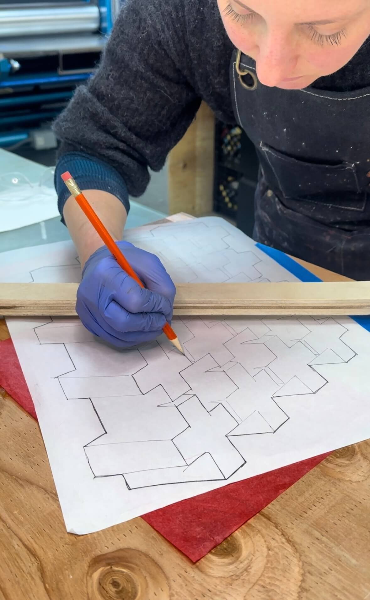 An artist in a black apron sketching a complex geometric pattern in pencil while wearing a purple glove, working at a wooden table.
