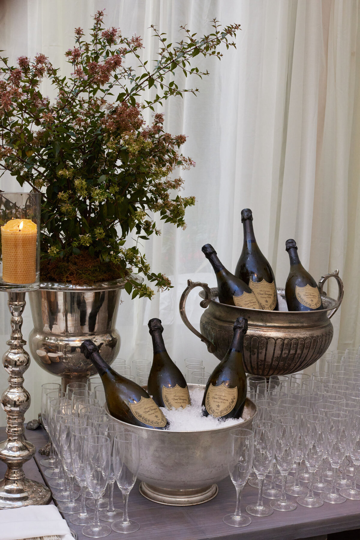 A collection of champagne bottles sits in three silver ice buckets on a table covered with empty champagne glasses. A tall candle burns in a silver holder beside the setup, and a lush floral arrangement in a silver vase is in the background.