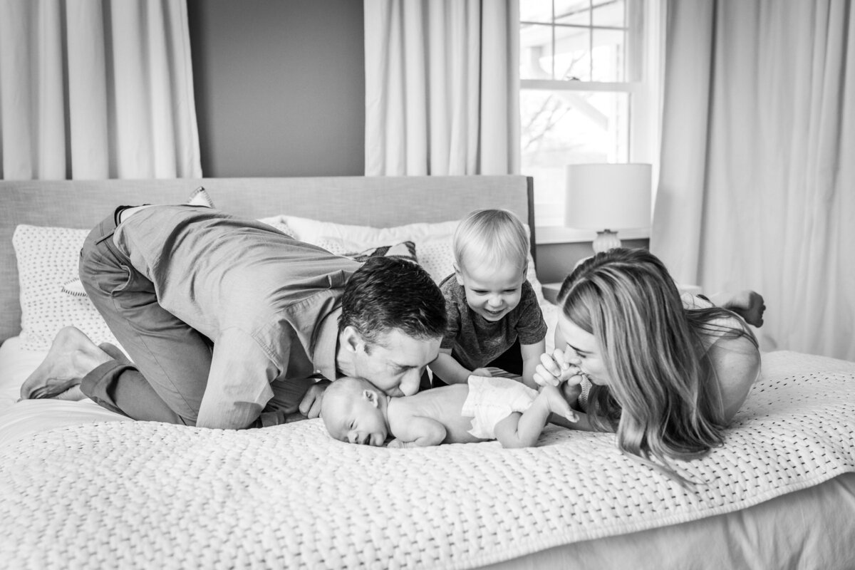 A black and white photo of a family of four lying on a bed, with the parents and toddler all leaning over to kiss and play with the newborn baby lying in the middle.