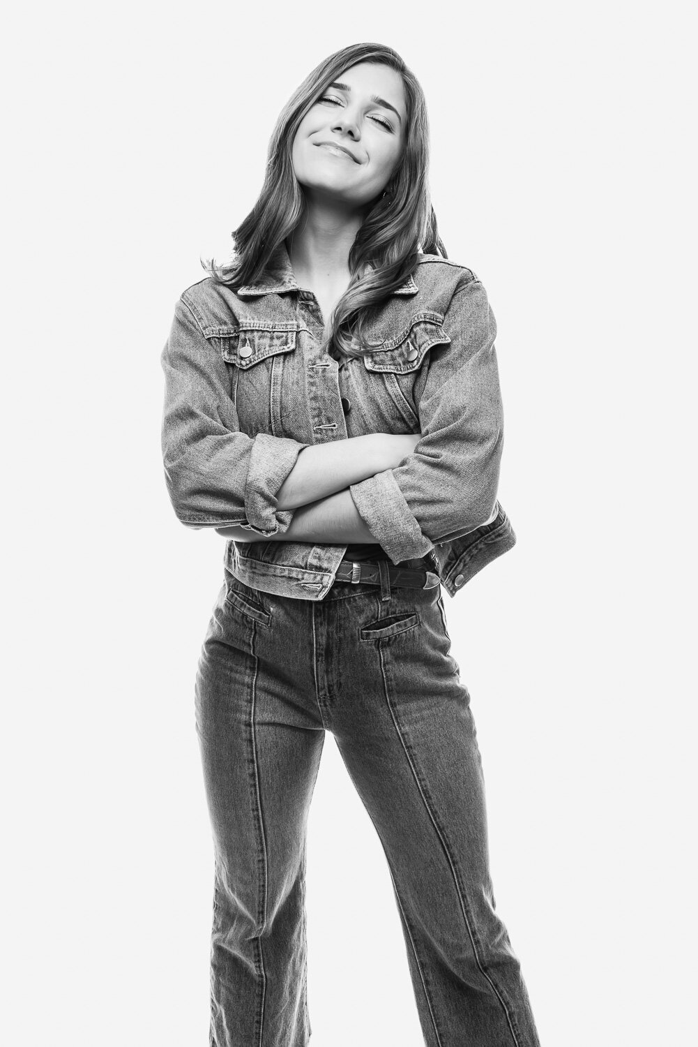Editorial-style high school senior portrait of a girl in blue jeans and a jean jacket, arms crossed and smiling blissfully, photographed on a white studio backdrop.