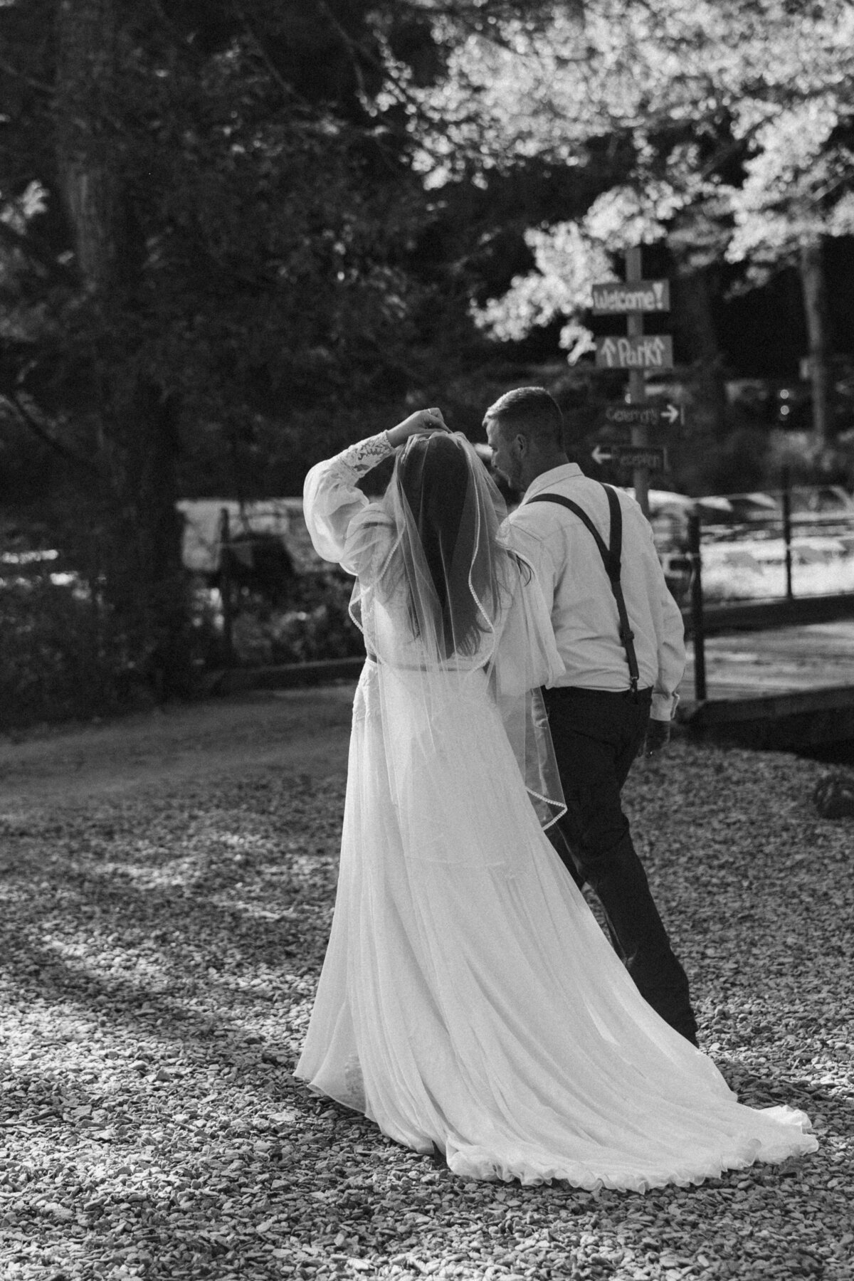 Black and white candid photo of a bride and groom walking hand in hand on a gravel path, with the bride lifting her veil slightly as sunlight filters through the trees.