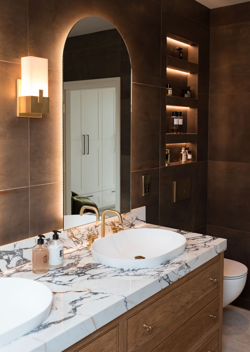 A modern bathroom with a marble countertop and wooden vanity featuring multiple drawers. The wall has dark, large tiles, and there's a gold faucet on the sink. Above the sink, an arched mirror reflects a door. To the left of the sink, sits a white toilet. Above the toilet, lit shelves are built into the wall, emitting a soft light.