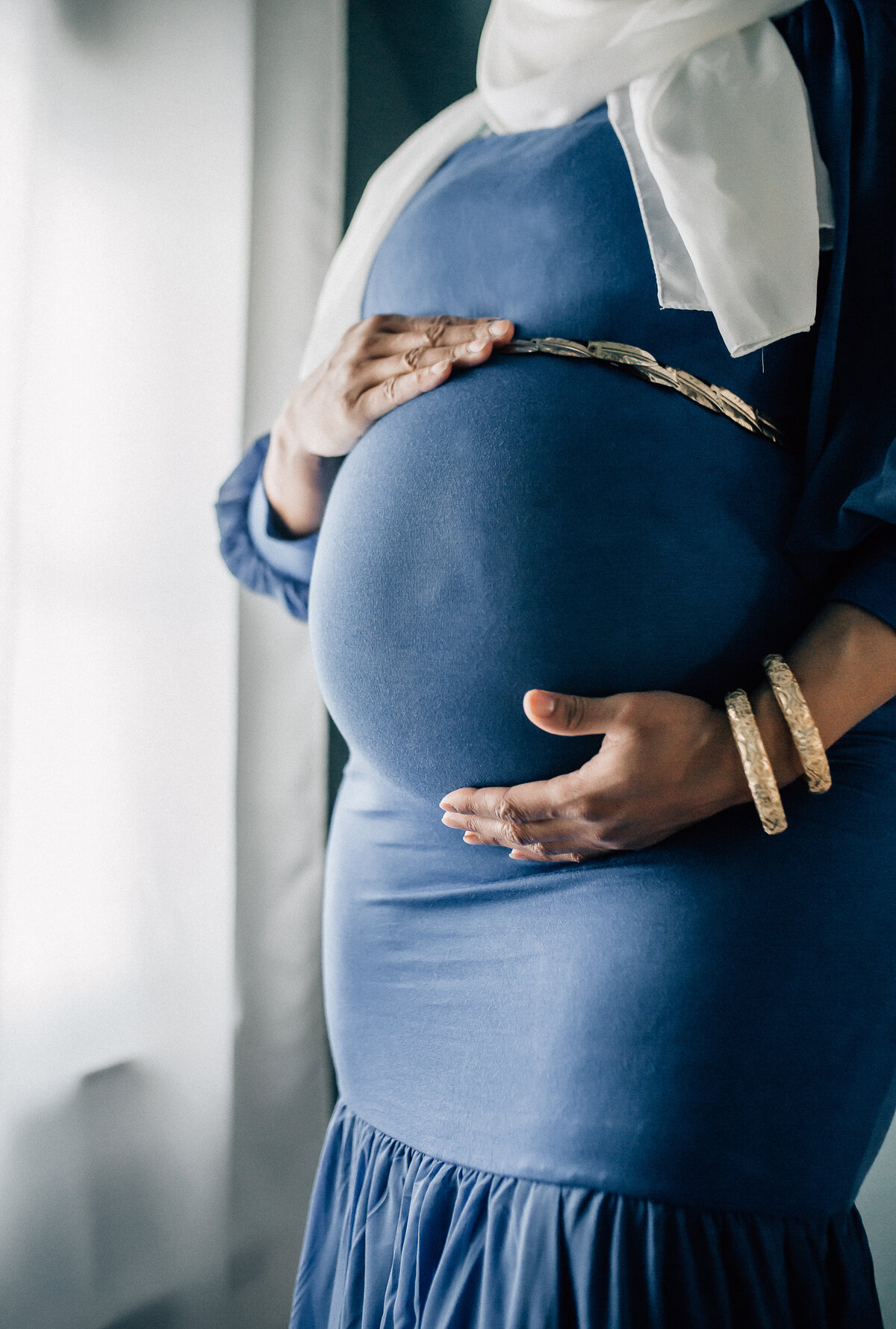 Maternity photoshoot with woman wearing blue dress, white scarf, and holding her belly.