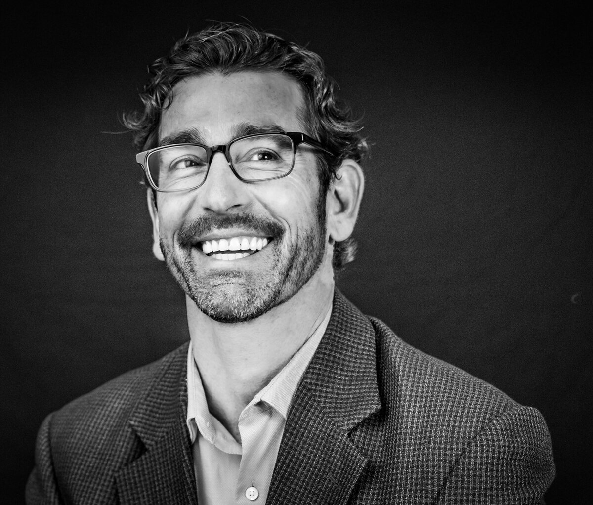 Black and white portrait of a bearded man wearing glasses and a jacket smiling at someone off camera