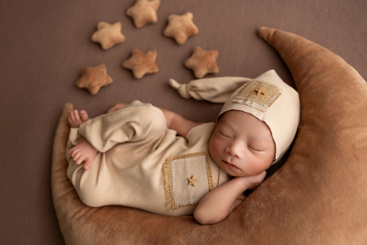 Newborn baby dressed in a beige outfit with a matching hat, sleeping on a crescent moon-shaped pillow, surrounded by small star pillows on a brown background.