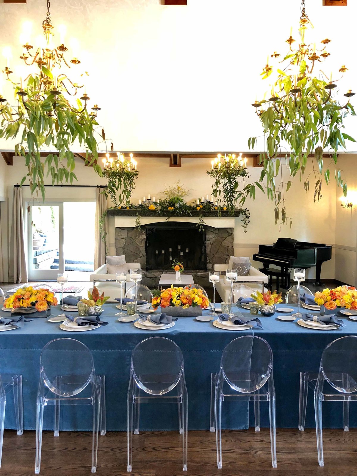 company dinner with orange centerpieces, blue linens, floating candles, clear lucite ghost chairs and greenery chandeliers