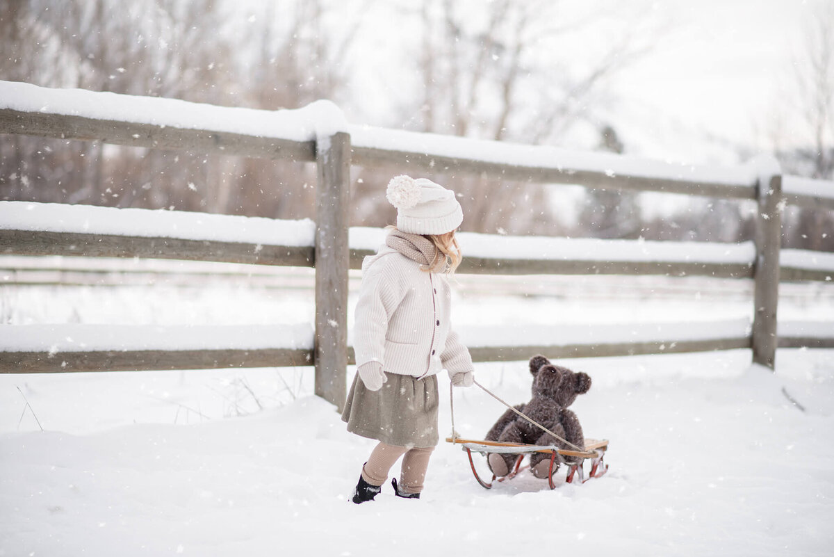 Little girl dressed in vintage winter layers pulls a teddy bear on a wooden sled through the snow. Captured in Kamloops by Brittany Danielle.