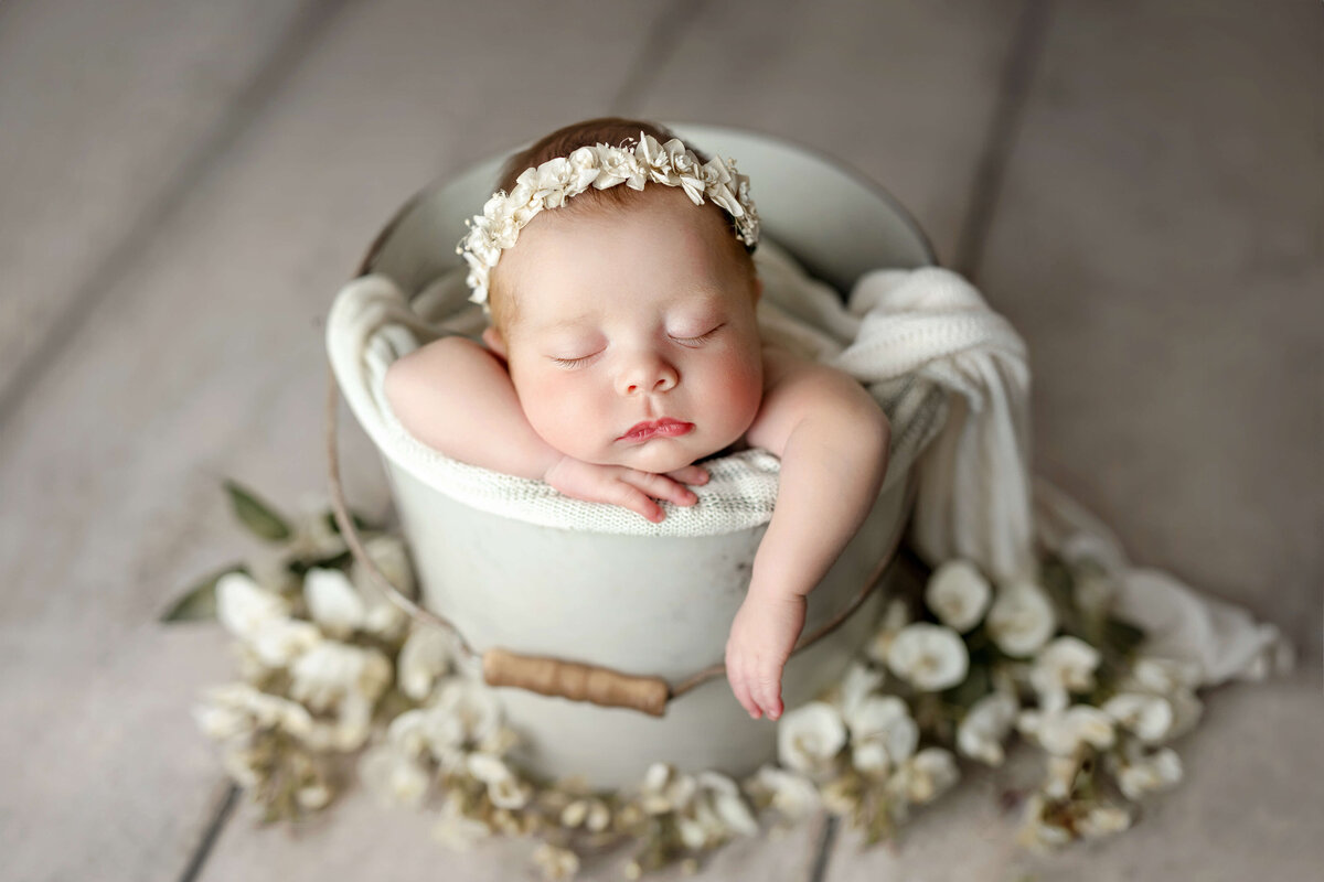 newborn baby girl laying in a white bucket with flowers around her with floral headband at a newborn photo shoot in Northern VA