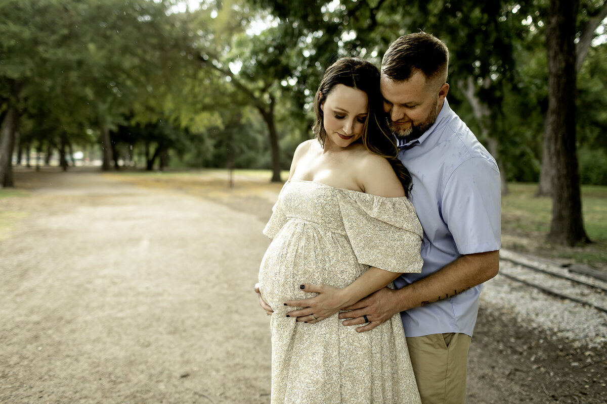 A loving maternity photo of mom and dad standing under a tree at Trinity Park, with dad gently resting his hand on mom’s belly, captured by Chunky Monkey Photography.
