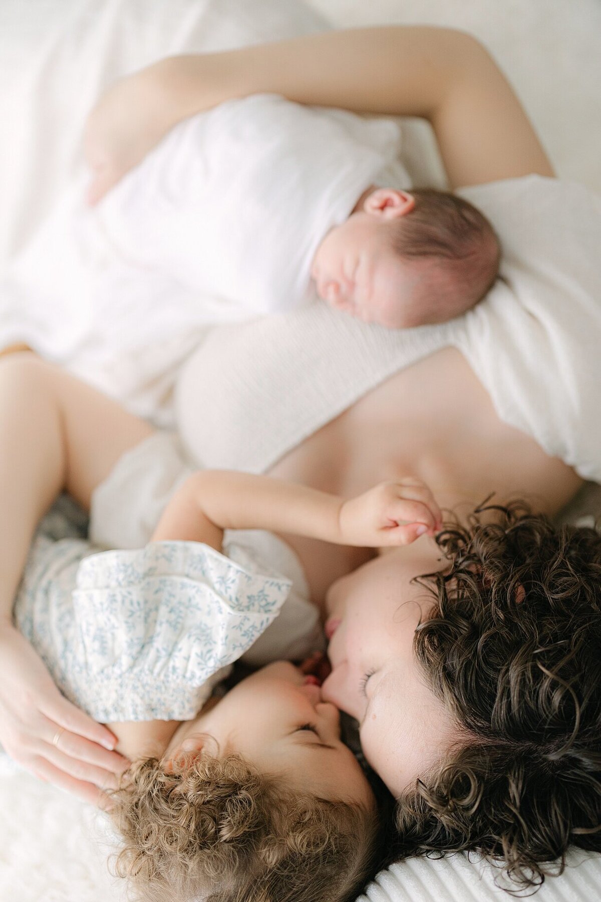 Mother lays holding her baby swaddled in white while snuggling with her toddler in a sage floral dress as she kisses her nose during a session with an Indianapolis newborn photographer