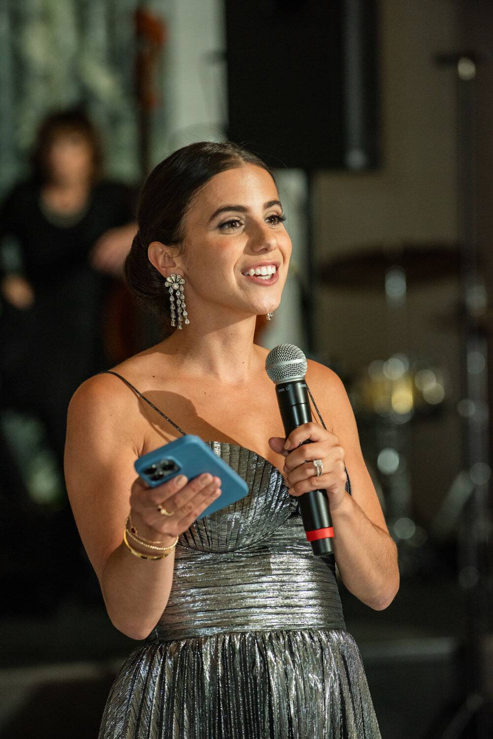 A woman in a silver dress is holding a microphone and a cell phone
