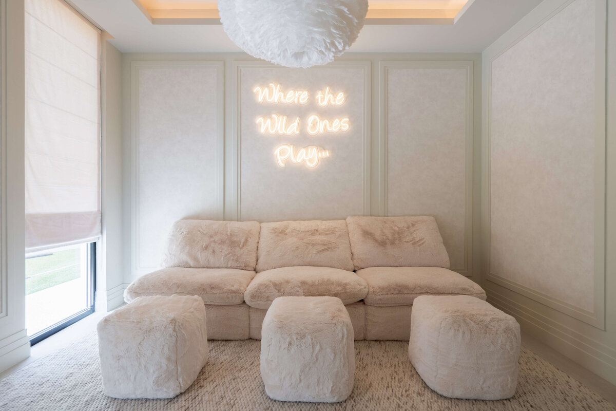 Spacious playroom with a light, airy aesthetic. There is a large, plush tan couch with three fuzzy pouf ottomans. Behind the couch is a softly illuminated neon  sign reading 'Where the Wild Ones Play...'. Above are four white, feathery spherical pendant chandeliers.