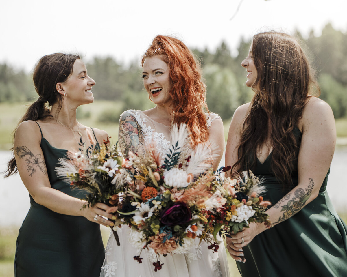 Three joyful women sharing a moment, with one in a white dress flanked by two in dark green, all holding rustic bouquets in an outdoor setting taken by jen Jarmuzek photography a Minneapolis wedding photographer