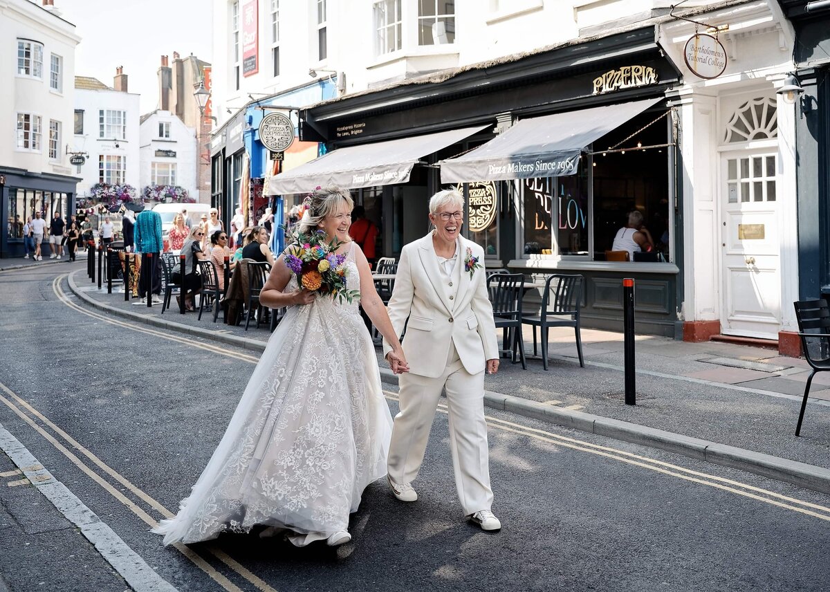 Two brides at a gay wedding holding hands and walking down the street laughing in Brighton
