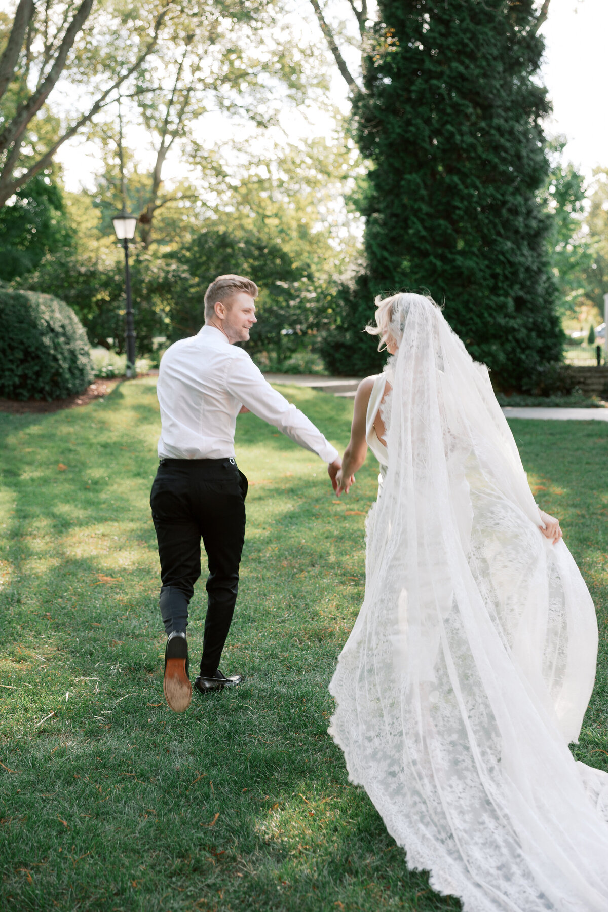 Groom in dress shirt and bride in veil run across lawn, candid wedding moment in Dayton, Ohio, by Philadelphia photographer.