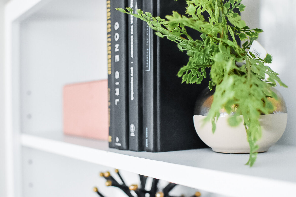 A small vase with greenery sits on a white bookshelf next to a stack of black books