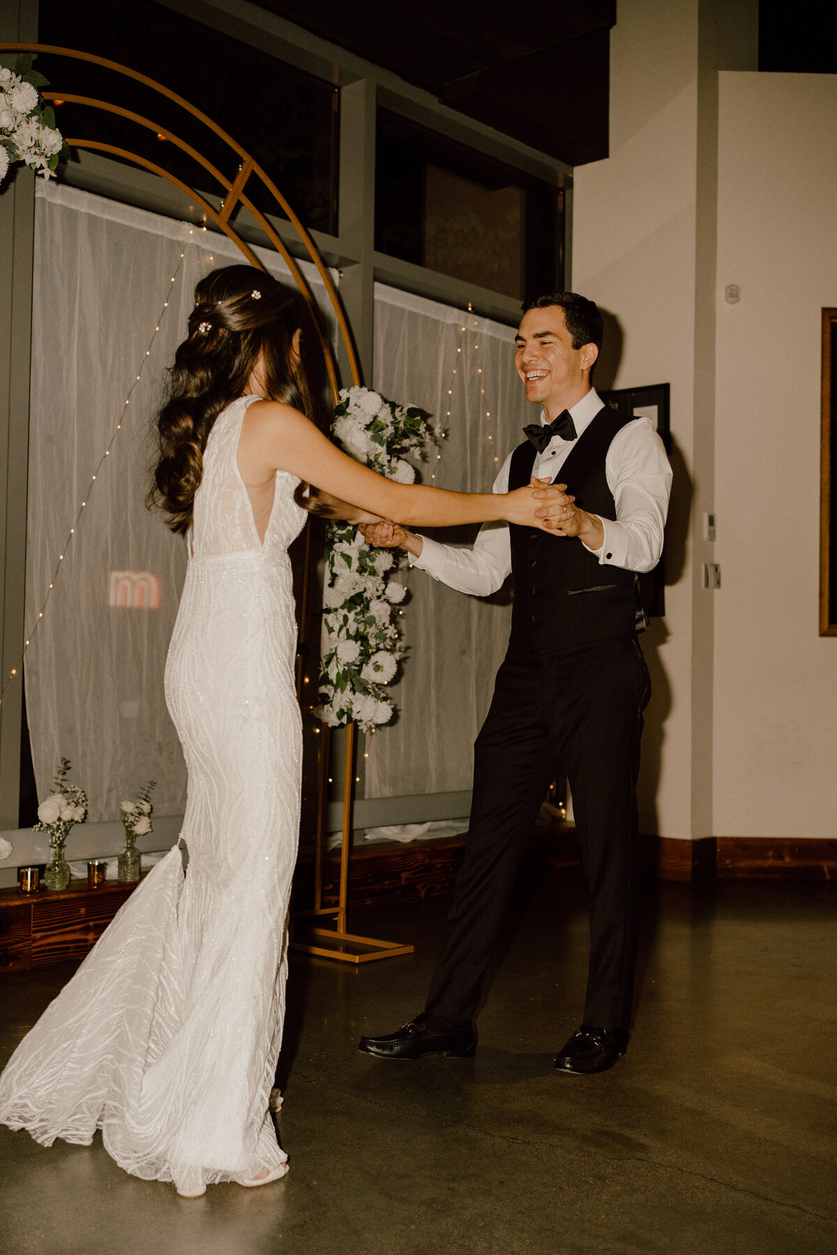 Bride and groom share a joyful dance at their wedding reception, with the bride in a stunning fitted lace gown and the groom in a stylish black tuxedo and vest. They dance in front of a beautifully decorated arch adorned with white flowers and twinkling lights, capturing a candid moment of happiness and celebration.