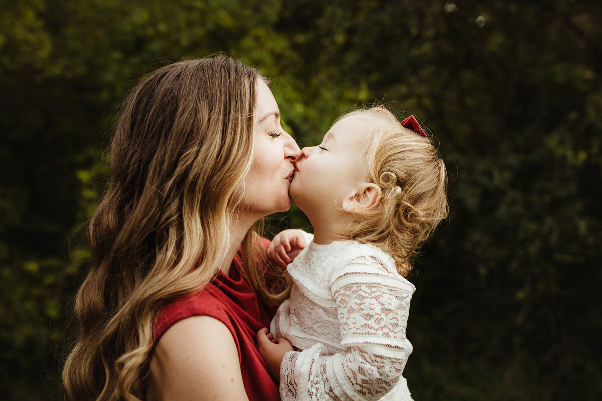 A beautiful mom and daughter kiss each other in front of the forest.