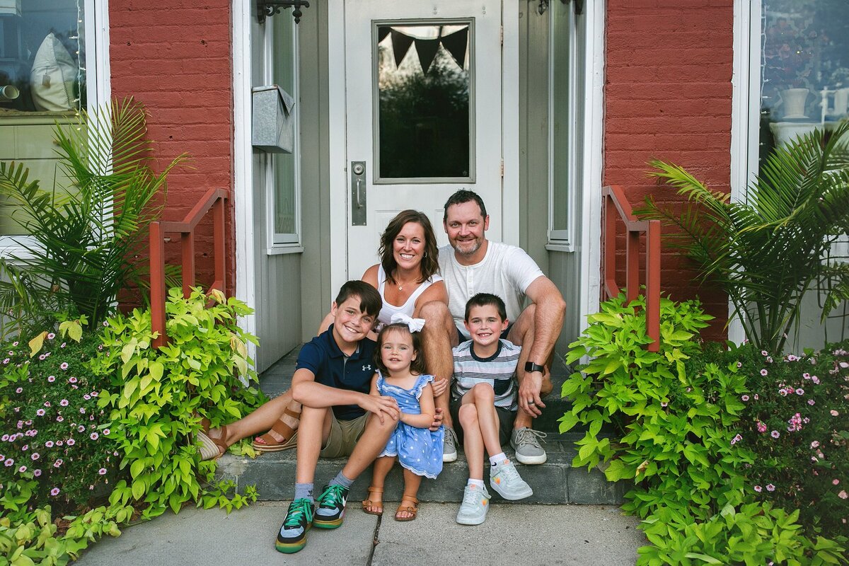 Smiling-family-of-five-sitting-on-front-porch-surrounded-by-greenery-during-a-relaxed-outdoor-photoshoot-in-zionsville-quaint-smalltown-main-street-vibe-family-photoshoot