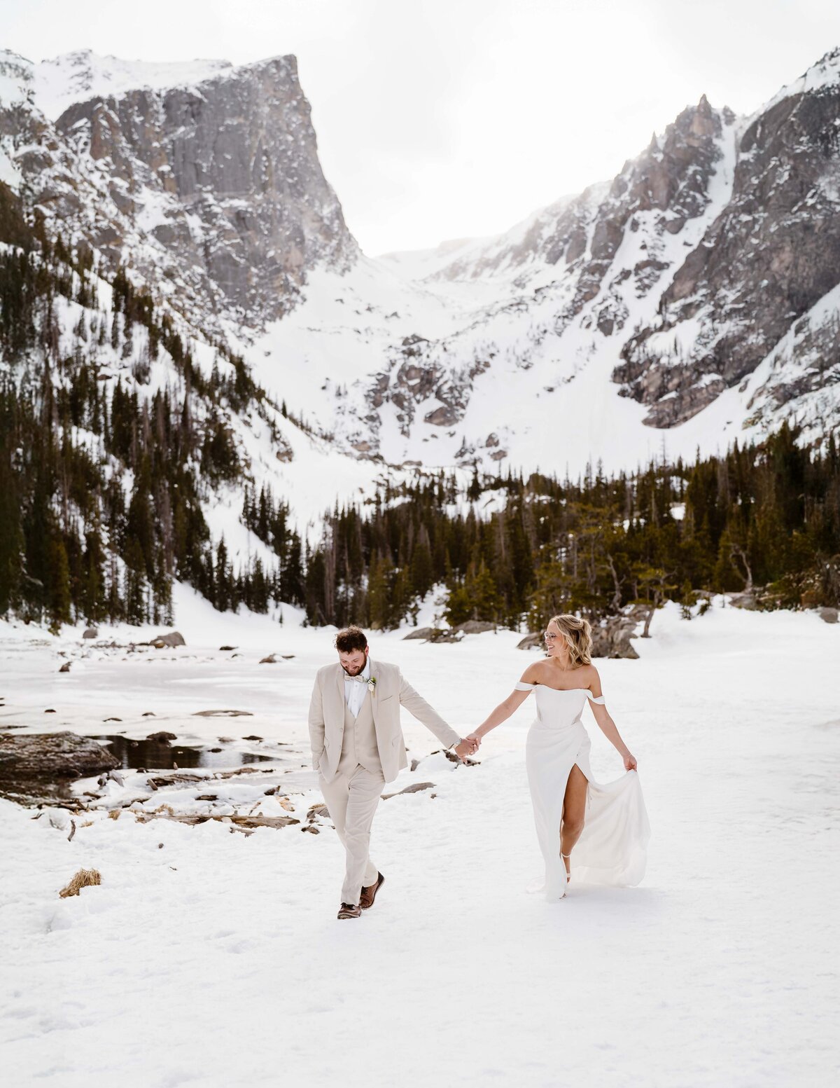 Couple stands hand in hand on the snowy shoreline of Bear Lake with mountain peaks rising behind them in Rocky Mountain National Park.