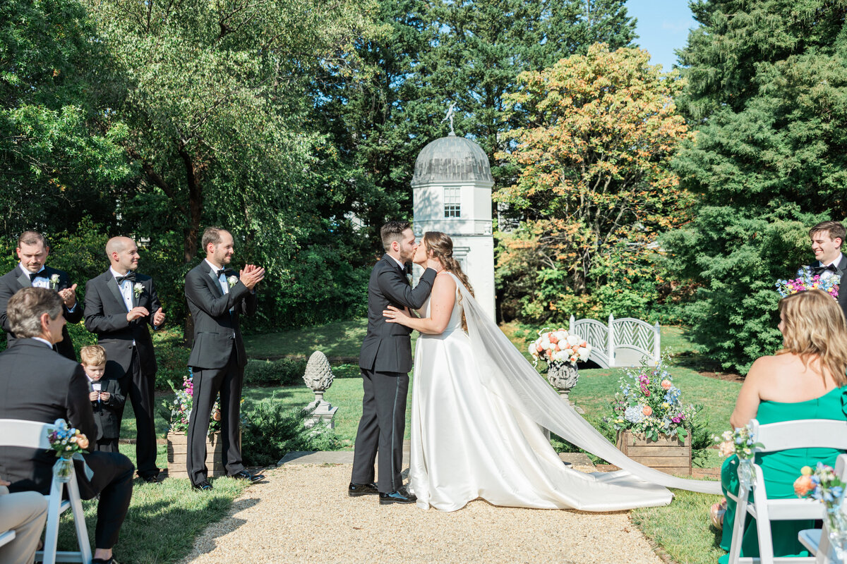 A couple during their first kiss at their wedding with guests cheering around them