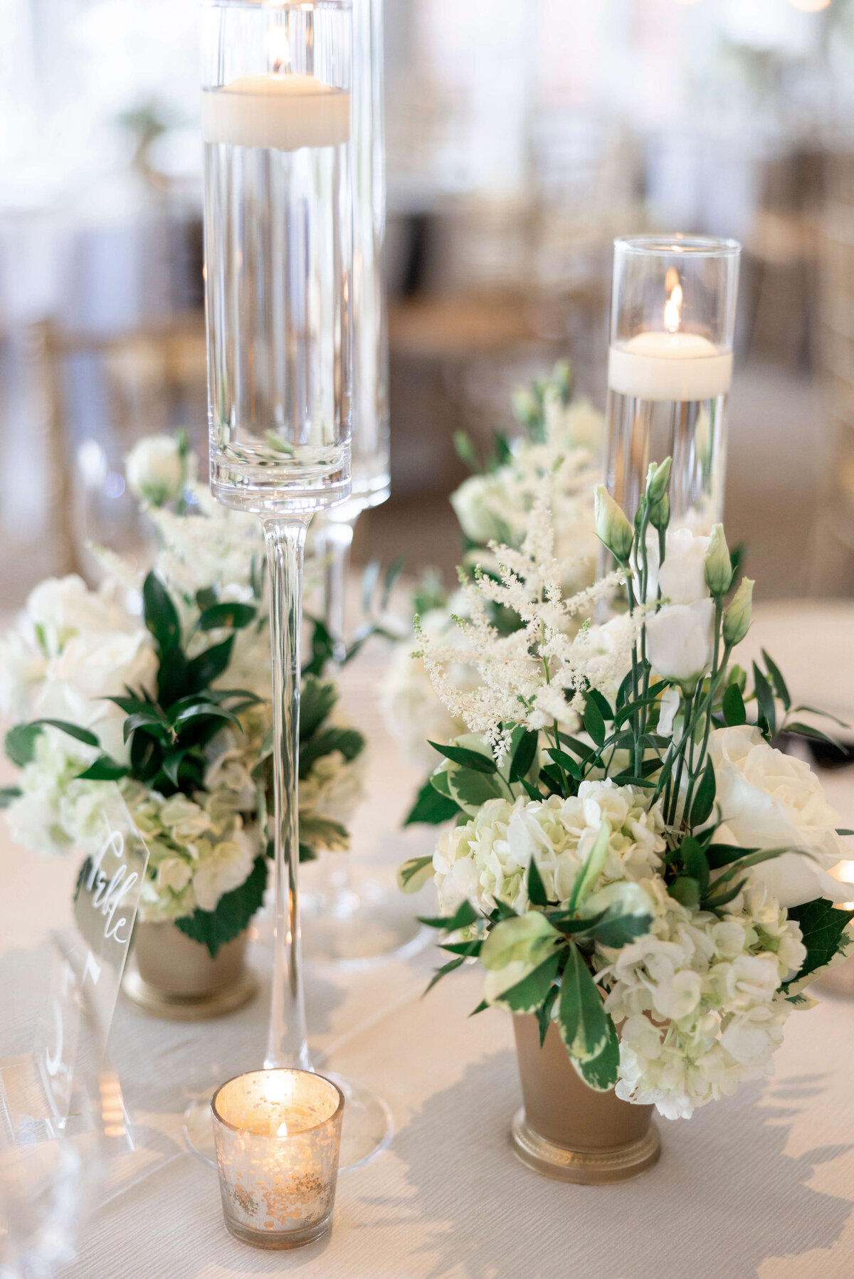 A wedding table centerpiece featuring elegant glass candle holders with lit candles and floral arrangements of white flowers and greenery. The table is softly lit, creating a romantic ambiance.