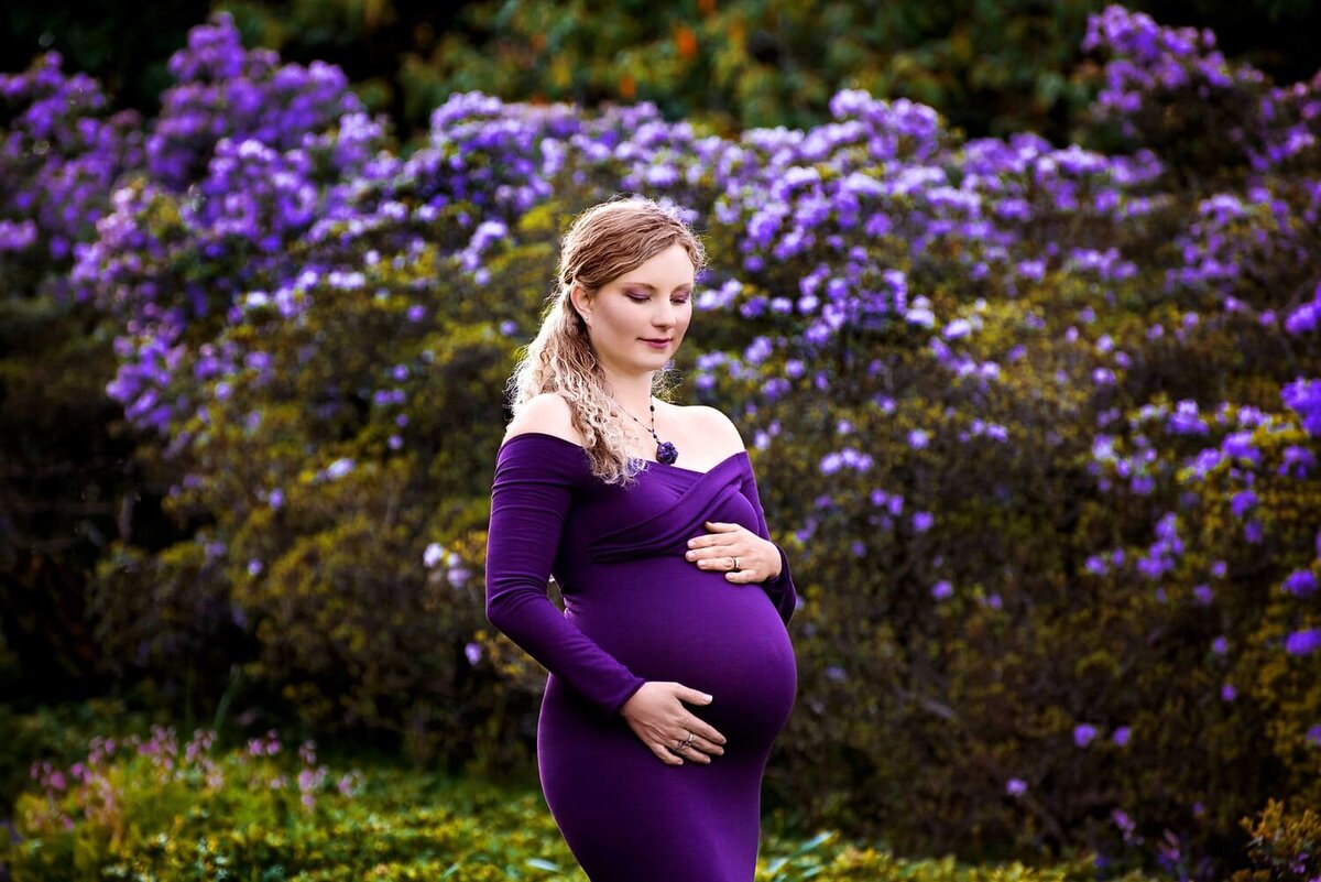 Mother to be in an amethyst purple maternity gown surrounded by flowers at Deer Lake Park in Burnaby