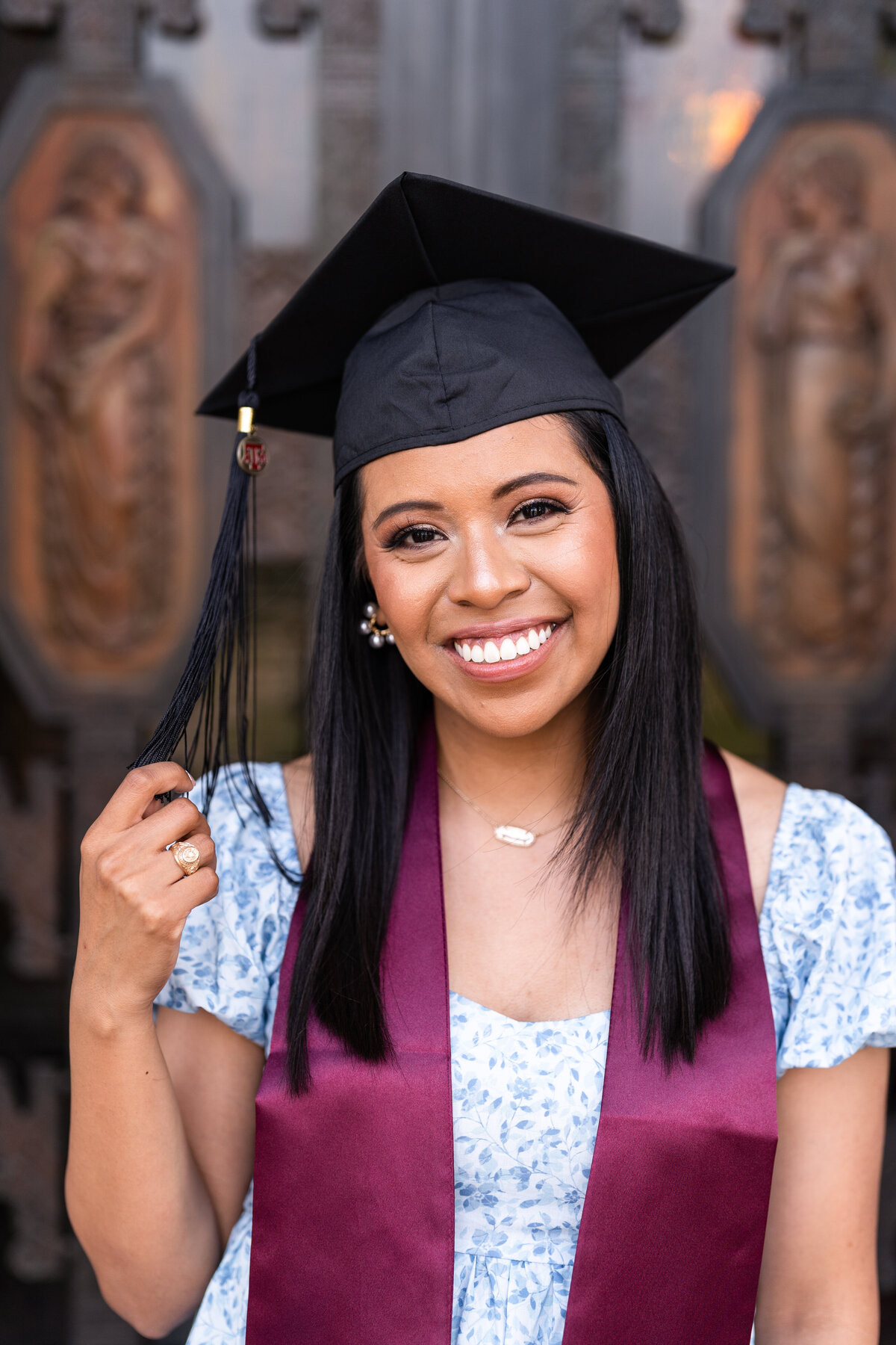 Texas A&M senior girl wearing cap and smiling while holding tassel and wearing maroon stole and blue dress in front of Administration building front doors