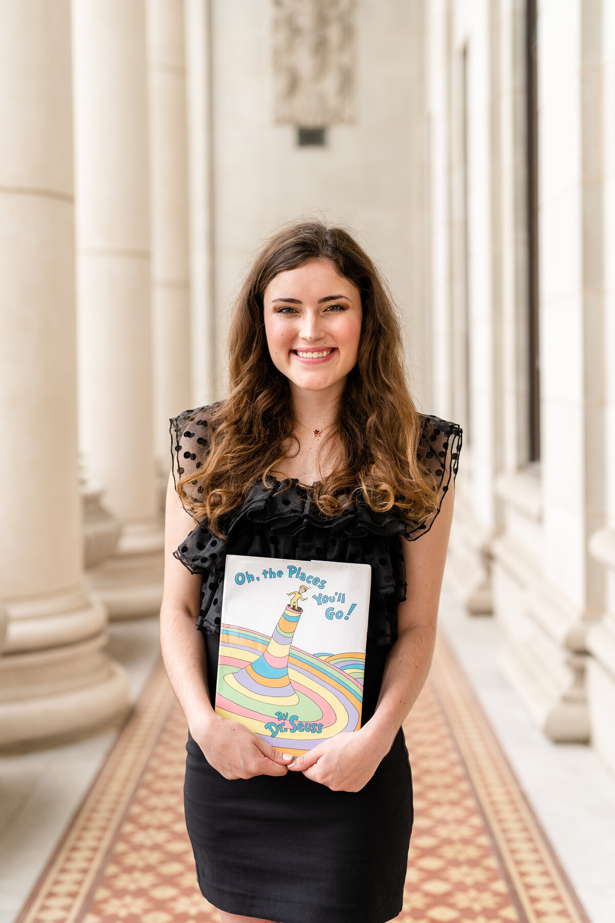 Texas A&M senior girl holding childhood book and smiling while wearing black dress in columns of Administration Building