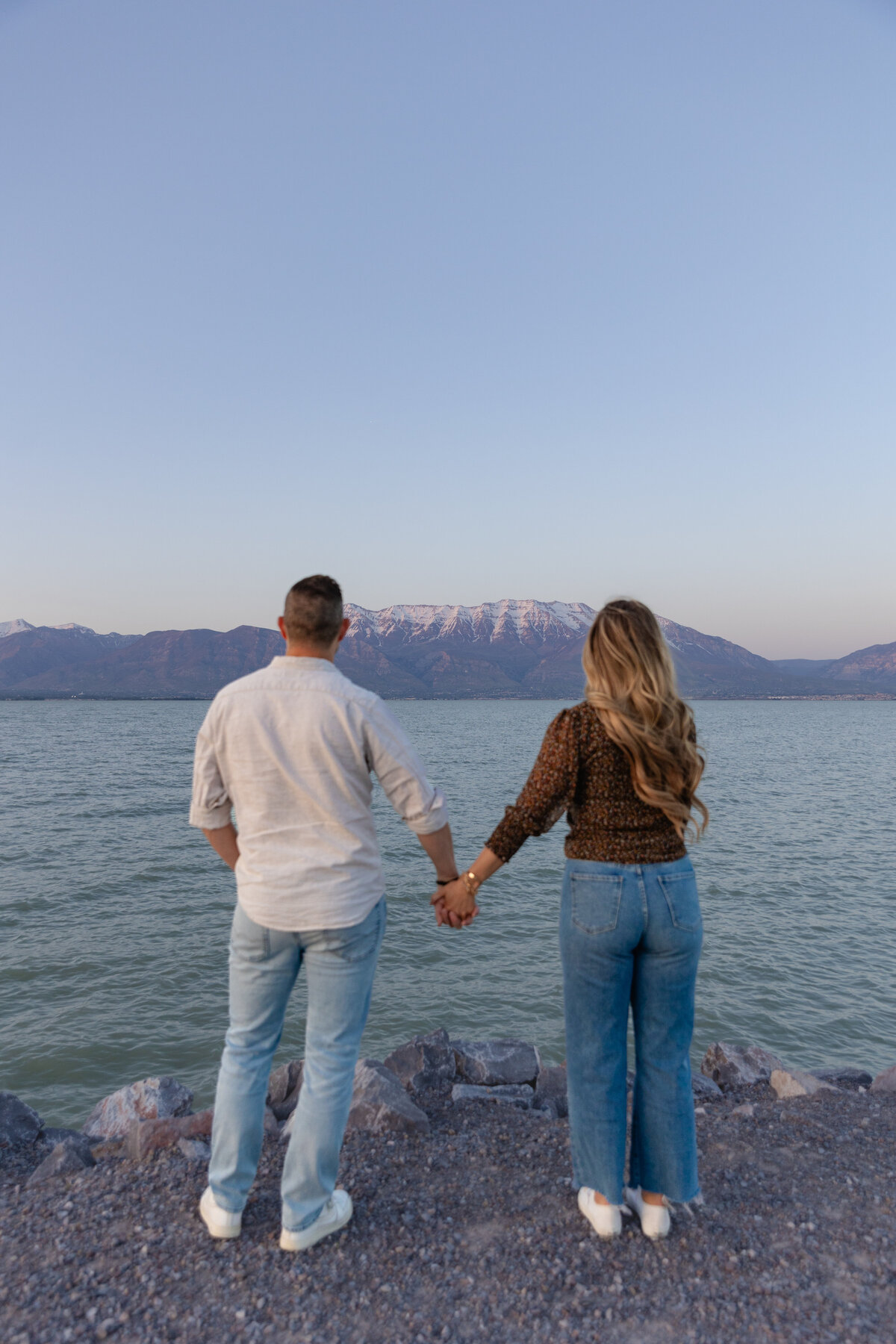 A couple stands hand in hand on the rocky shoreline of Utah Lake in Saratoga Springs, UT, gazing at the snow-capped mountains in the distance. The evening sky casts a soft glow over the water.