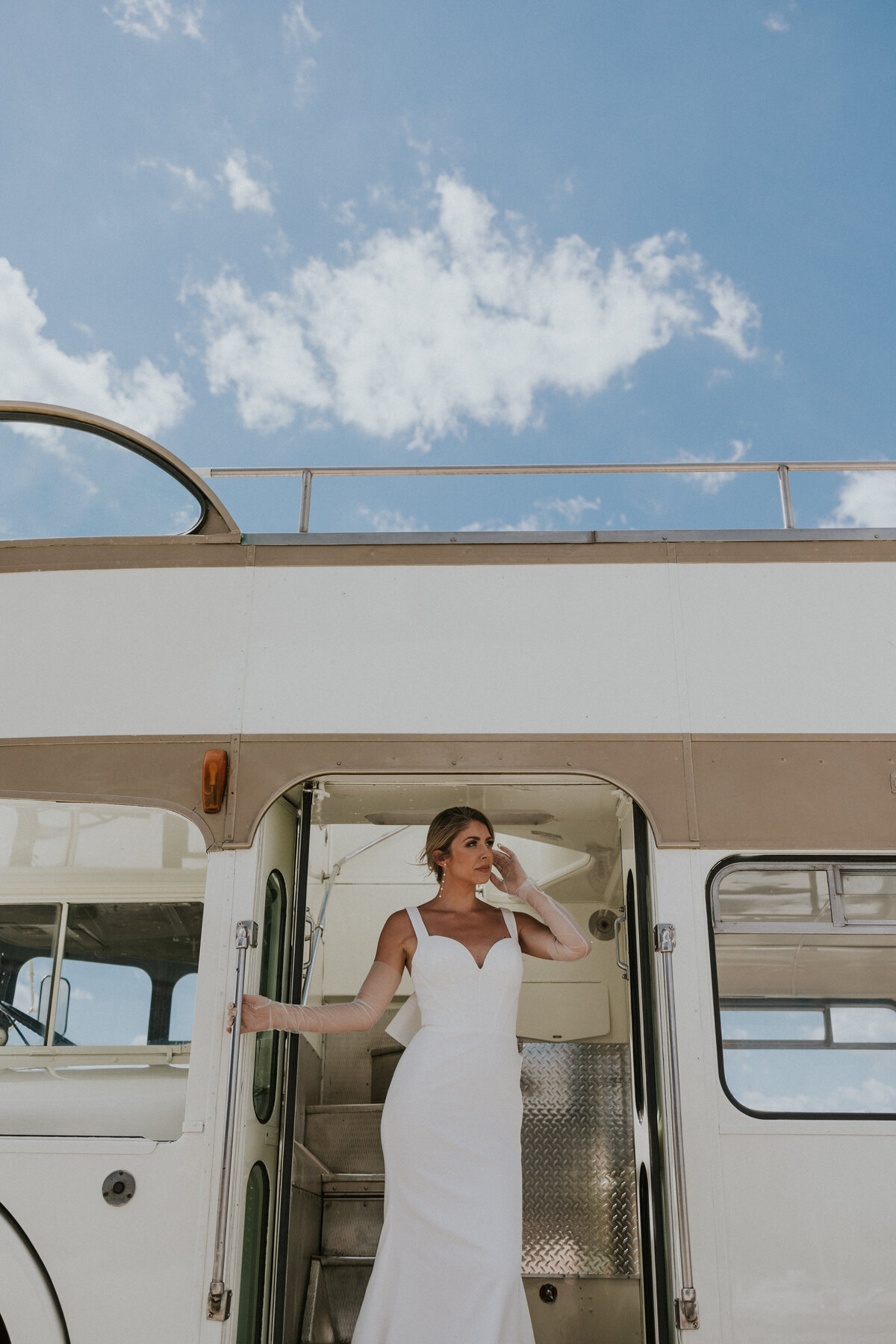 Bride stands at the doorway of double decker bus while cloud floats by in the sky at the top of the frame.